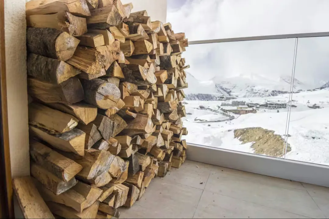 Bois de chauffage rangé devant la baie vitrée de Maison Georgia, la neige sur les pentes au-dehors